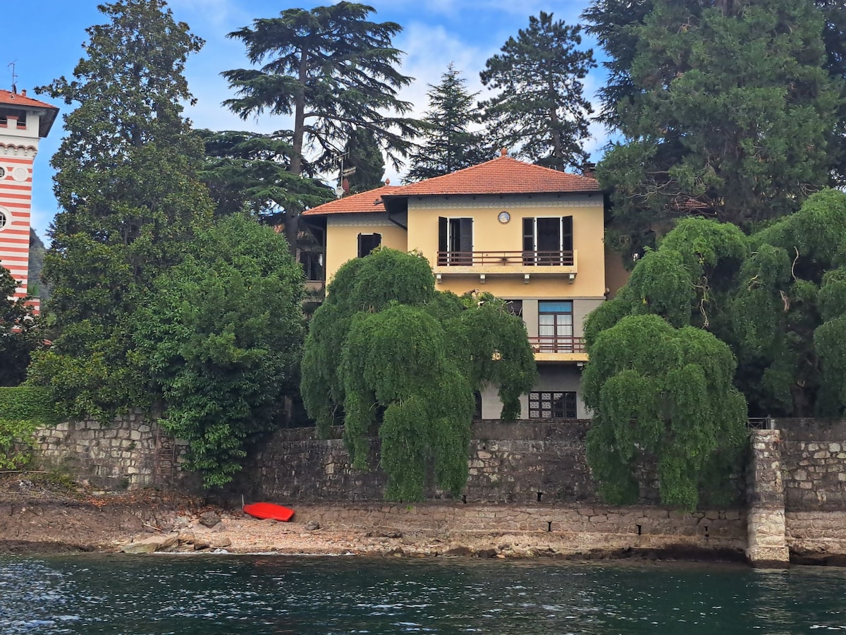 The three-story house features a balcony overlooking the lake, with a sloping roof and large windows. Lush greenery surrounds the property, providing shade. A small red kayak is visible on the shore, complementing the natural setting by the water.