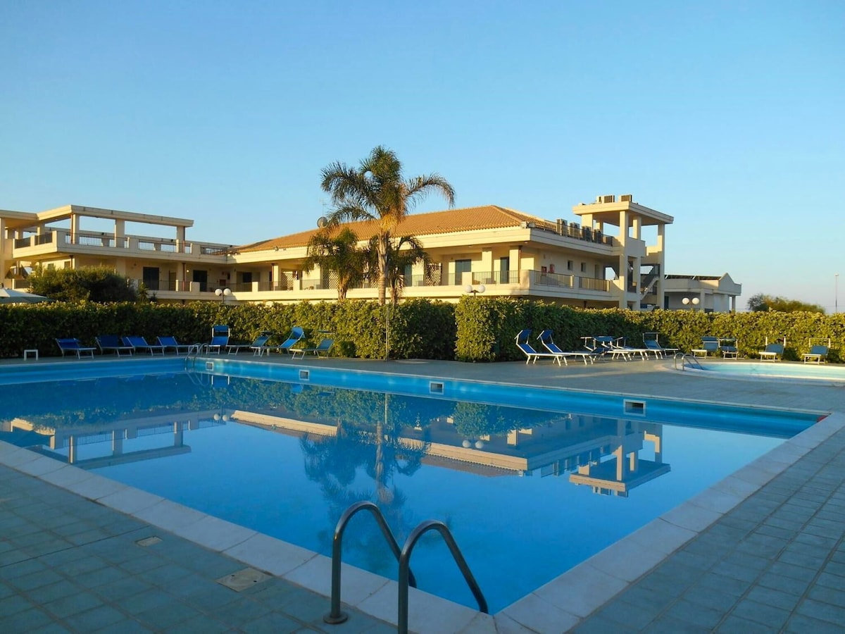 A swimming pool is surrounded by lush greenery, reflecting the clear blue sky. Sun loungers line the poolside, providing ample space for relaxation. The multi-level building in the background complements the serene outdoor environment.