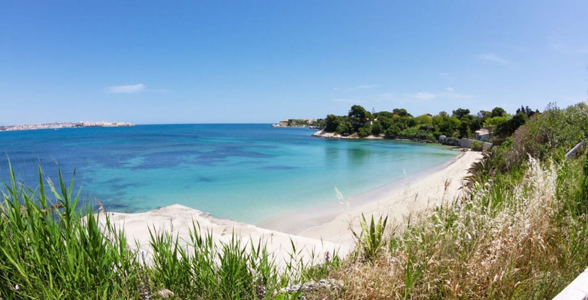 A serene beach scene is displayed, featuring soft sand and calm turquoise waters. Gentle waves lap at the shore, while greenery lines the beach, creating a peaceful transition between land and sea. The clear sky enhances the tranquil atmosphere of this coastal setting.