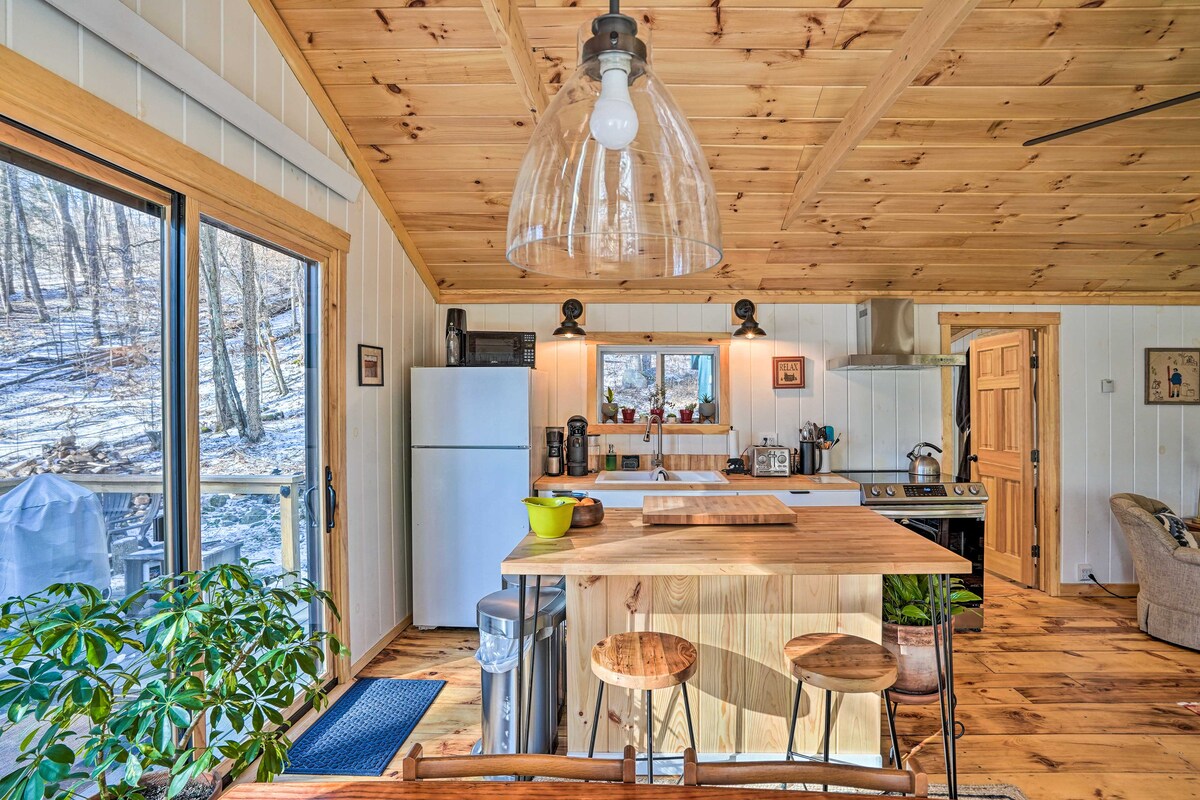 An open kitchen space features a wood-topped island with seating for two. A refrigerator and stove are positioned against the wall, with window views of the surrounding woods. Natural light floods the area, highlighting the wood paneling and rustic decor of the cabin.