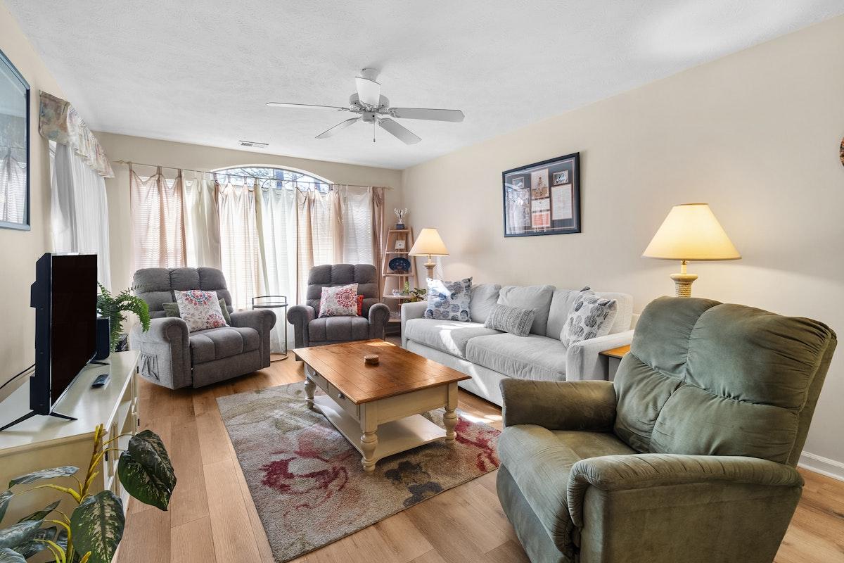 A spacious living area features soft seating, including two gray recliners and a light-colored sofa. A wooden coffee table occupies the center, surrounded by a decorative area rug. Natural light enters through large windows dressed with sheer curtains, creating a bright and inviting atmosphere.