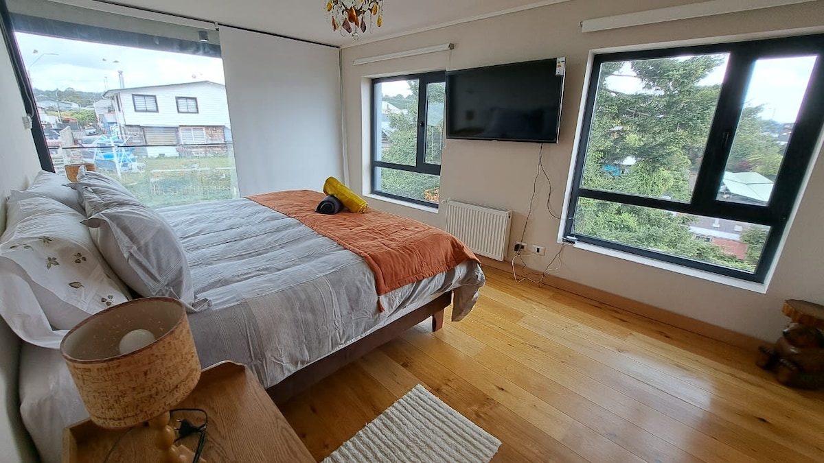 A well-lit bedroom features a spacious bed with neutral bedding and an orange accent blanket. Large windows allow for ample natural light and offer views of the outside. A television is mounted on the wall, and warm wooden flooring contributes to the inviting space.