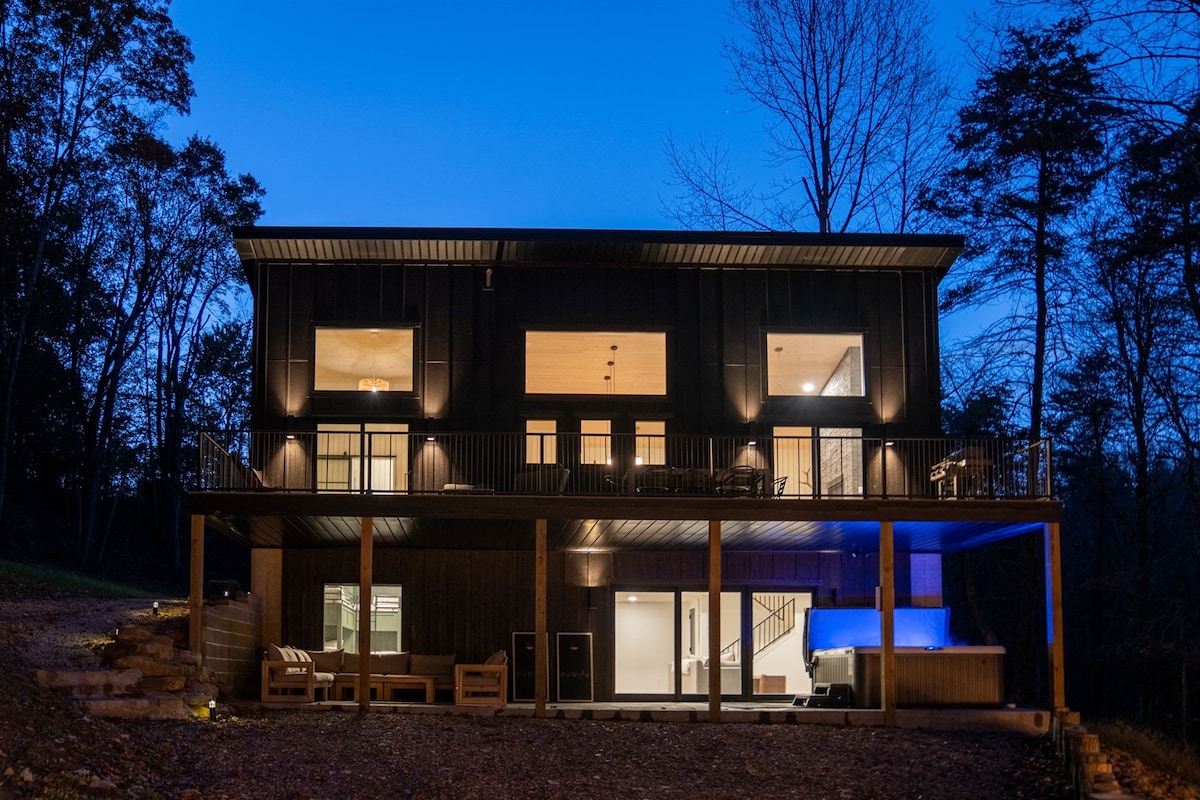 The exterior of the cabin is displayed at twilight, showcasing a modern design with dark wood and metal accents. Soft lighting highlights the expansive windows, while the inviting covered patio features seating. The surrounding trees create a serene backdrop, enhancing the natural setting.