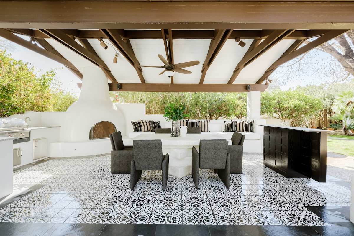 An outdoor dining area is featured under a shaded patio, showcasing a large table surrounded by comfortable chairs. The floor is elegantly tiled with contrasting black and white patterns. A charming fireplace and a well-equipped outdoor kitchen are positioned in the background, enhancing the culinary experience.
