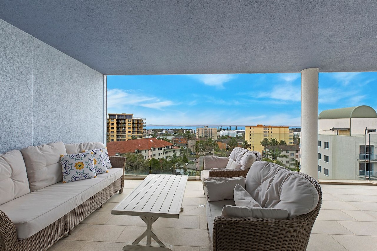 A spacious balcony is furnished with comfortable seating, featuring light-colored cushions on a white table, offering panoramic views of the surrounding area and the lake. The sky is partially cloudy, enhancing the setting's openness.