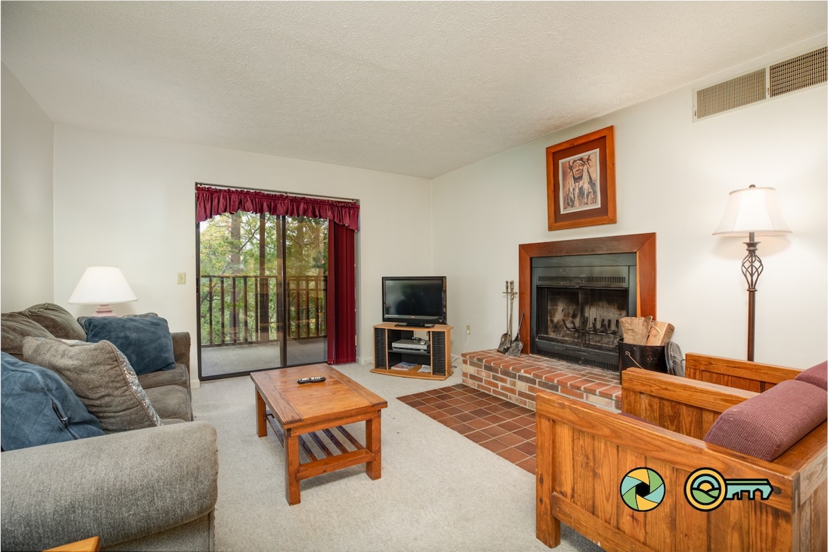 The living room is furnished with a gray sofa, a wooden armchair, and a coffee table. A flat screen TV is positioned near a wood-burning fireplace. Natural light enters through the sliding glass door, which leads to a balcony surrounded by trees.