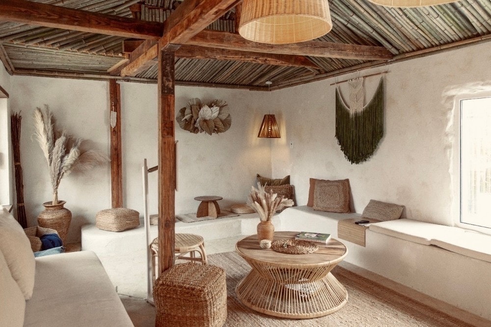 A cozy living space features natural wood beams and a textured ceiling, creating an inviting atmosphere. A round woven coffee table is surrounded by various seating options, including a light-colored sofa and several textured cushions. Soft natural light enters through a window, illuminating the warm decor.