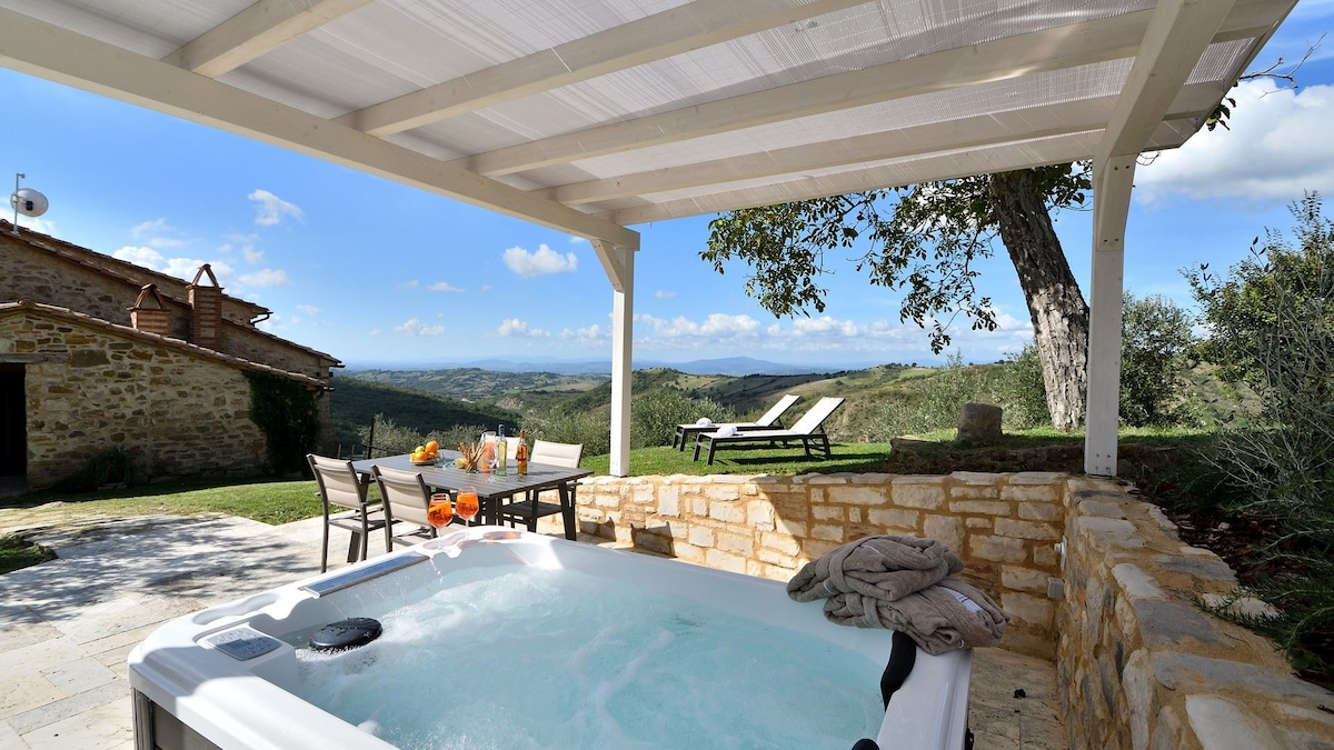 A private outdoor space features a Jacuzzi under a shaded pergola. A stone wall surrounds the area, and sun loungers are positioned nearby, offering views of the undulating Tuscan landscape. A dining table is set for al fresco meals, surrounded by greenery.