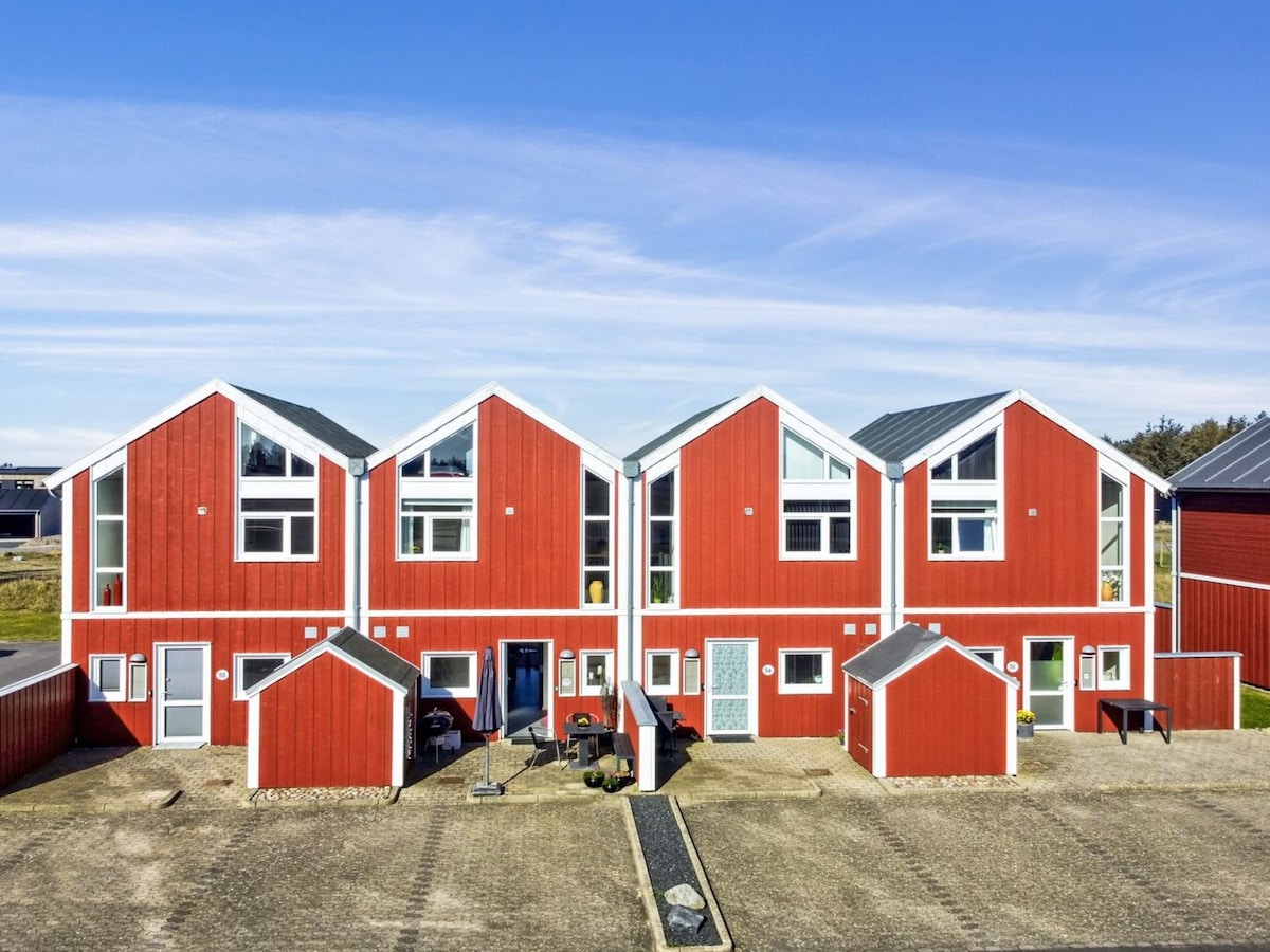 Colorful red houses with sloped roofs are arranged in a row, showcasing large windows on each unit. The pathway leads to the entrances of the houses, bordered by landscaped areas. Natural surroundings are visible in the background under a clear blue sky.