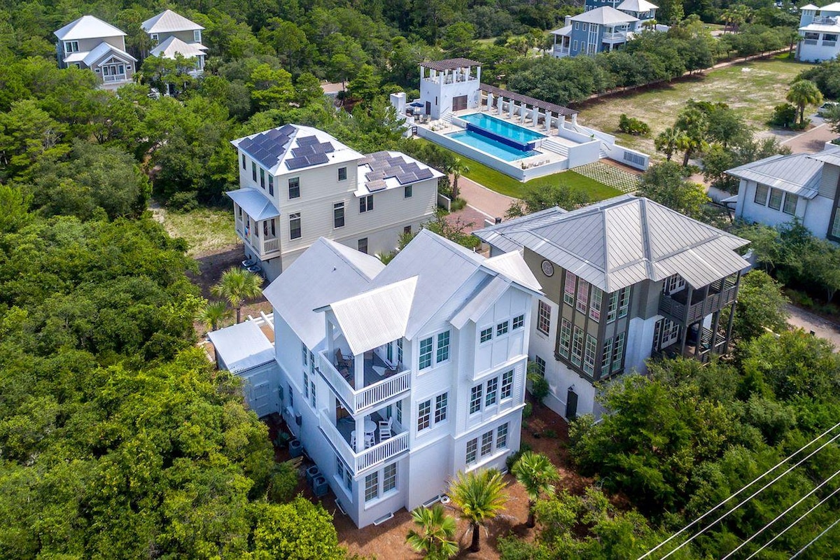 An aerial view showcases the contemporary home nestled among lush greenery within a gated community. The striking design features multiple levels and a large outdoor area. A community infinity pool is visible nearby, surrounded by lounge chairs and landscaping.