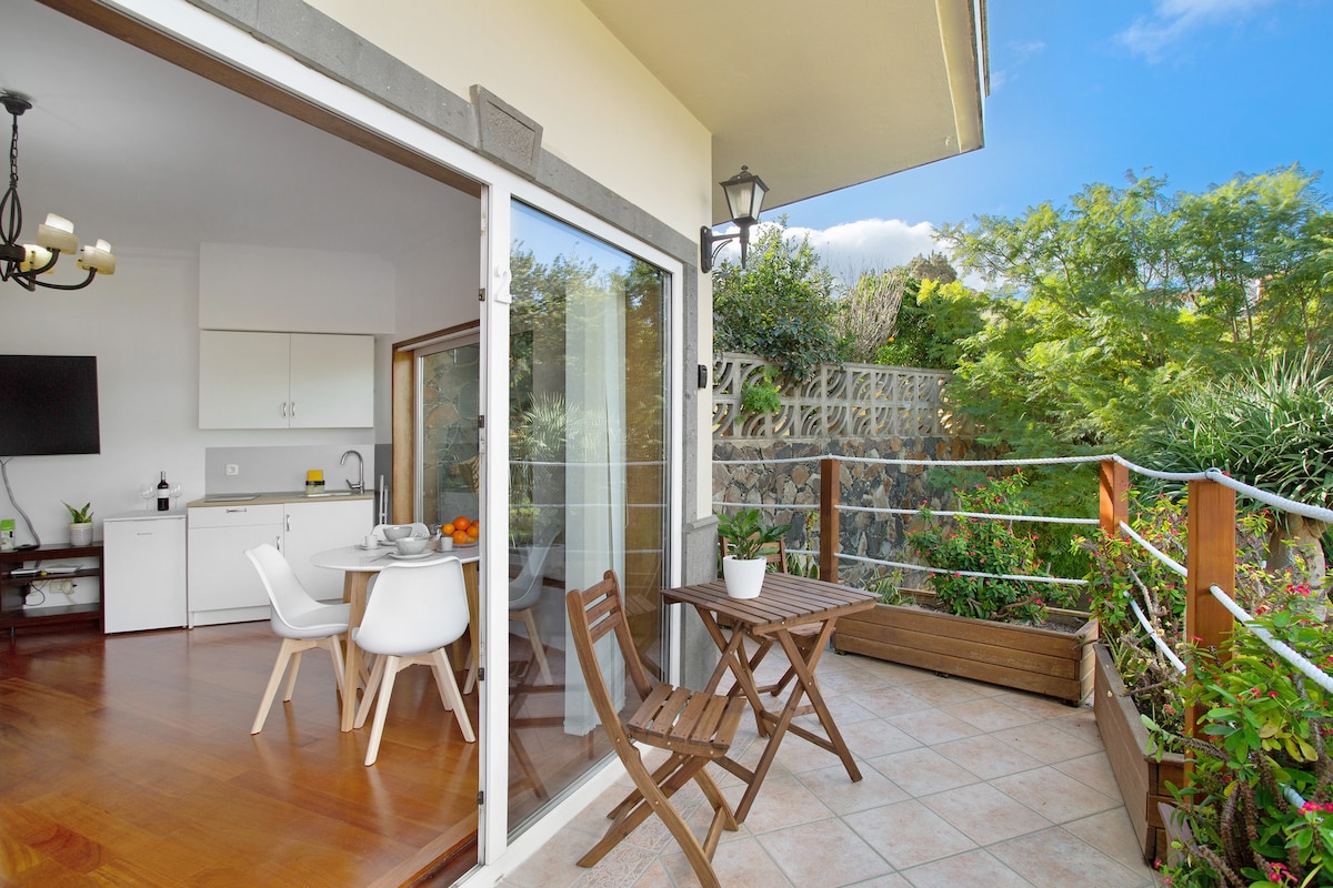 An open terrace is shown, featuring a wooden table and chairs, with a small potted plant on the table. The adjacent area reveals a well-lit kitchen and dining space through a sliding glass door, enhancing connectivity with the natural surroundings.
