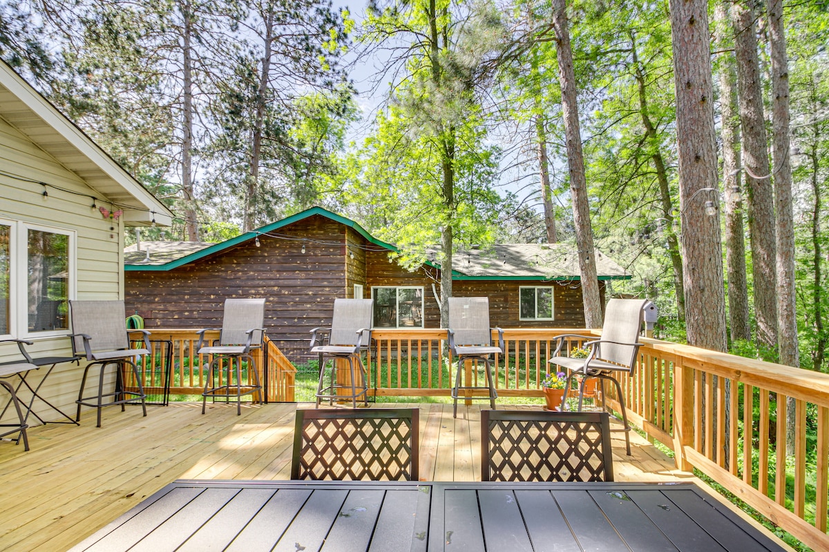 A spacious wooden deck is surrounded by tall trees, providing a natural setting. Several high-backed chairs face outward, ready for relaxation. A dining set is positioned slightly lower on the deck, offering additional seating options.