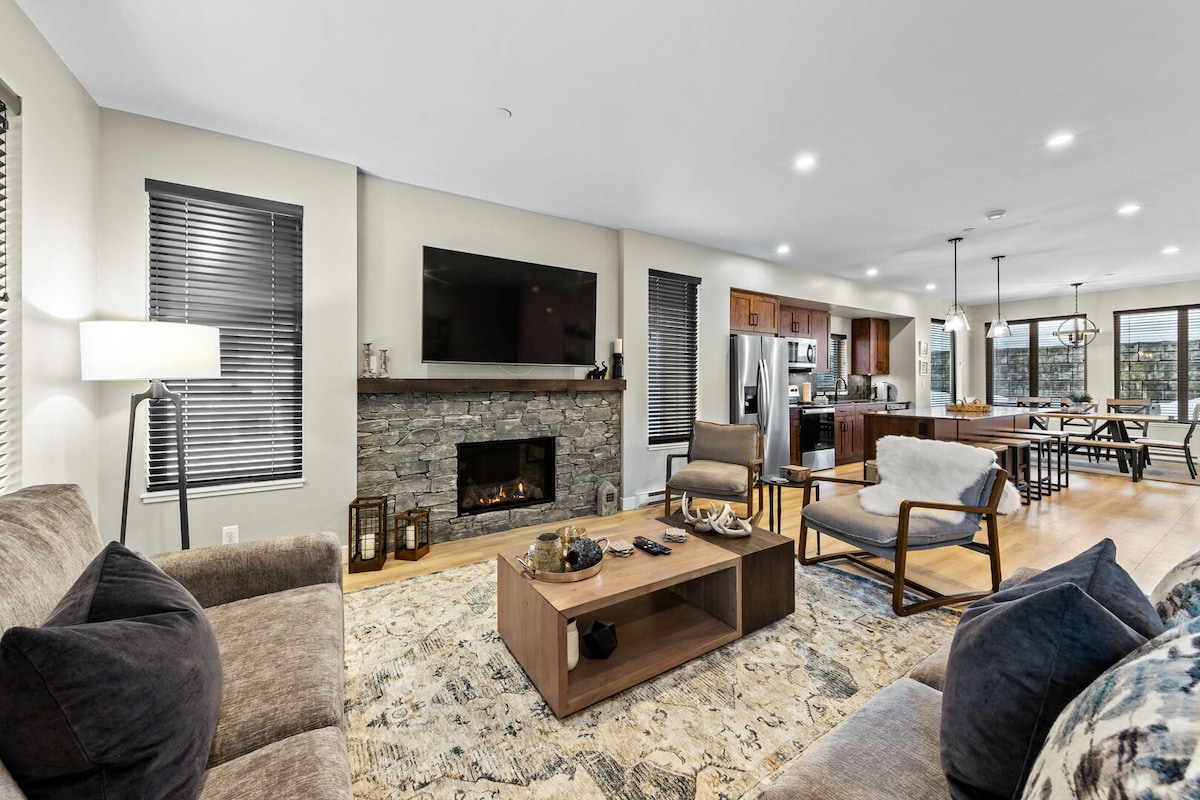 An inviting living area features comfortable seating options and a stylish coffee table. A stone fireplace serves as a focal point beneath a large flat-screen TV. Natural light is provided by multiple windows dressed with minimalist blinds, while the open layout connects to the kitchen and dining area.