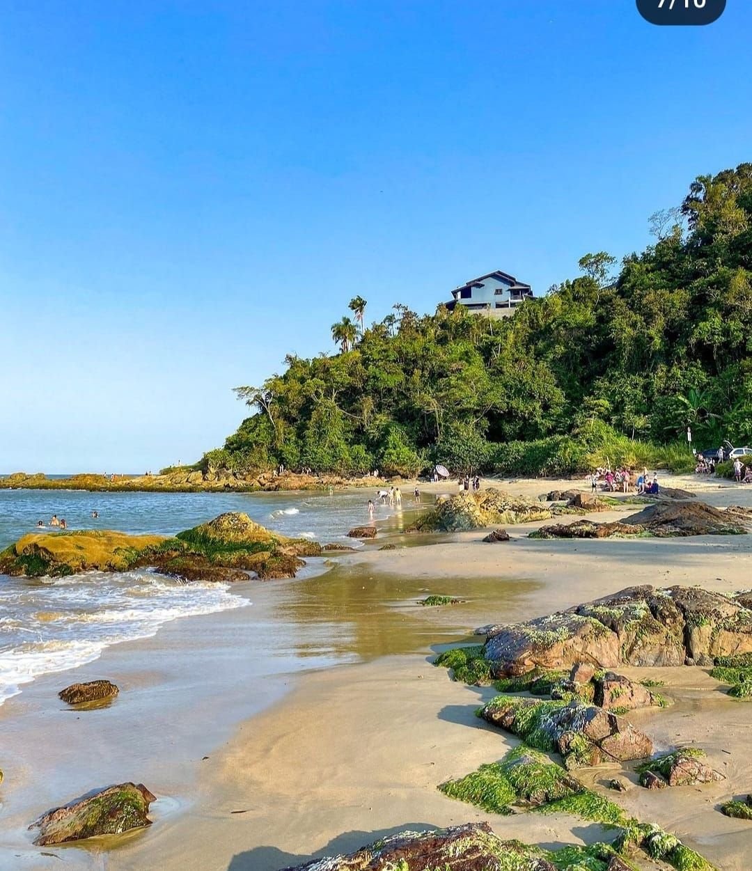 The image captures a serene beach scene with gentle waves lapping against the shore. Rocky formations and patches of greenery are visible along the coastline. A house is situated atop a hill, surrounded by trees. People can be seen enjoying the beach.