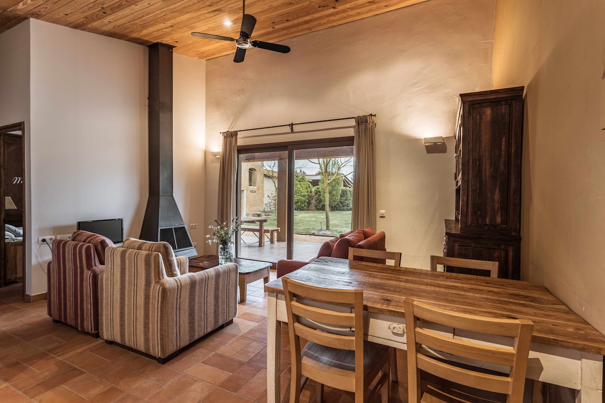 The living area features a wooden dining table surrounded by light-colored chairs. A comfortable seating arrangement of striped sofas is positioned near a large window, offering views of the outdoor space. A fireplace adds warmth to the room, complemented by wooden ceiling beams.