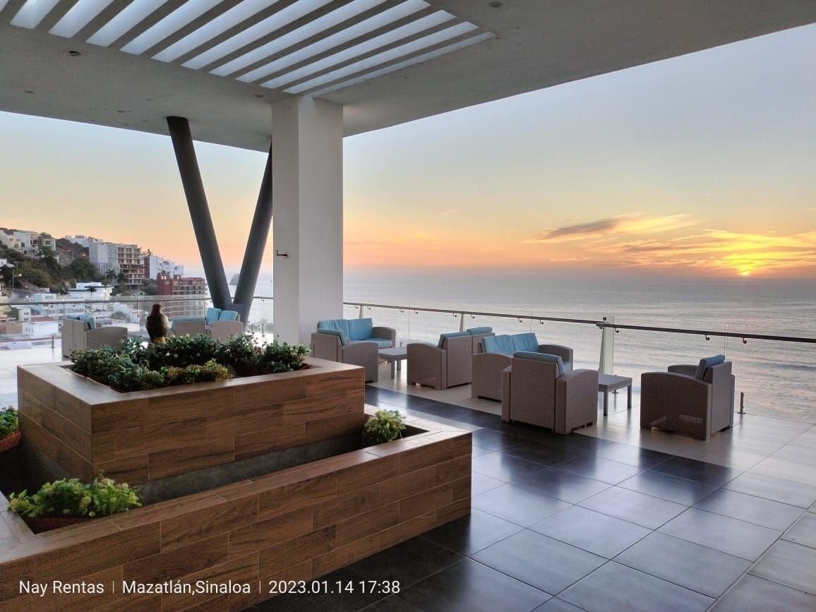 An outdoor terrace offers views of the ocean and sunset, furnished with seating arrangements in soft hues. Planters with greenery create a natural element in the space, while large glass panels provide a sense of openness and connection to the scenic surroundings.