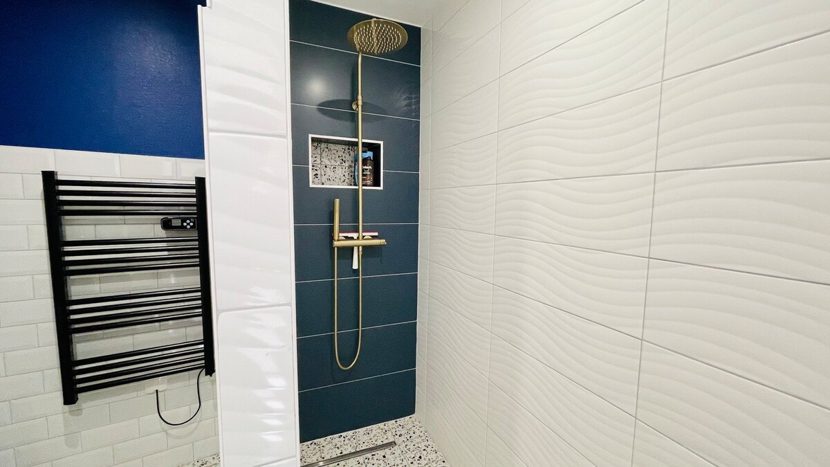 A spacious shower area is highlighted by elegant gold fixtures against a backdrop of textured tiles. The walls feature a mix of smooth white and deep blue tiles, with a small niche for toiletries. A heated towel rack is positioned nearby for convenience.