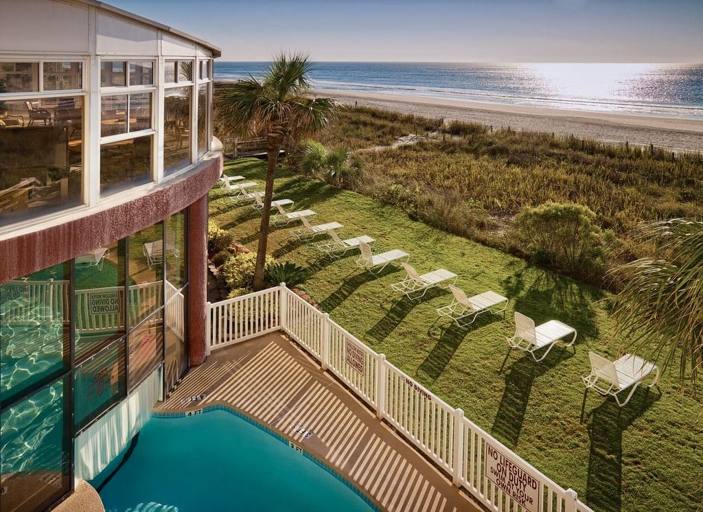 An outdoor area features a well-manicured lawn bordered by a path leading to the beach. Sun loungers are positioned on the grass, and a swimming pool is visible adjacent to the building, with sunlight casting shadows across the pool deck.