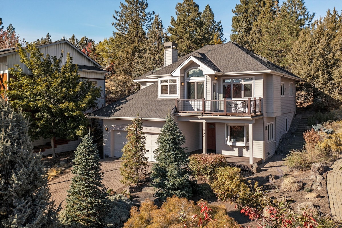 The exterior of the home is shown, featuring a light-colored facade and a prominent balcony over the entrance. Lush landscaping, including evergreen trees and colorful shrubs, surrounds the driveway leading to a two-car garage. The clear blue sky enhances the inviting atmosphere.