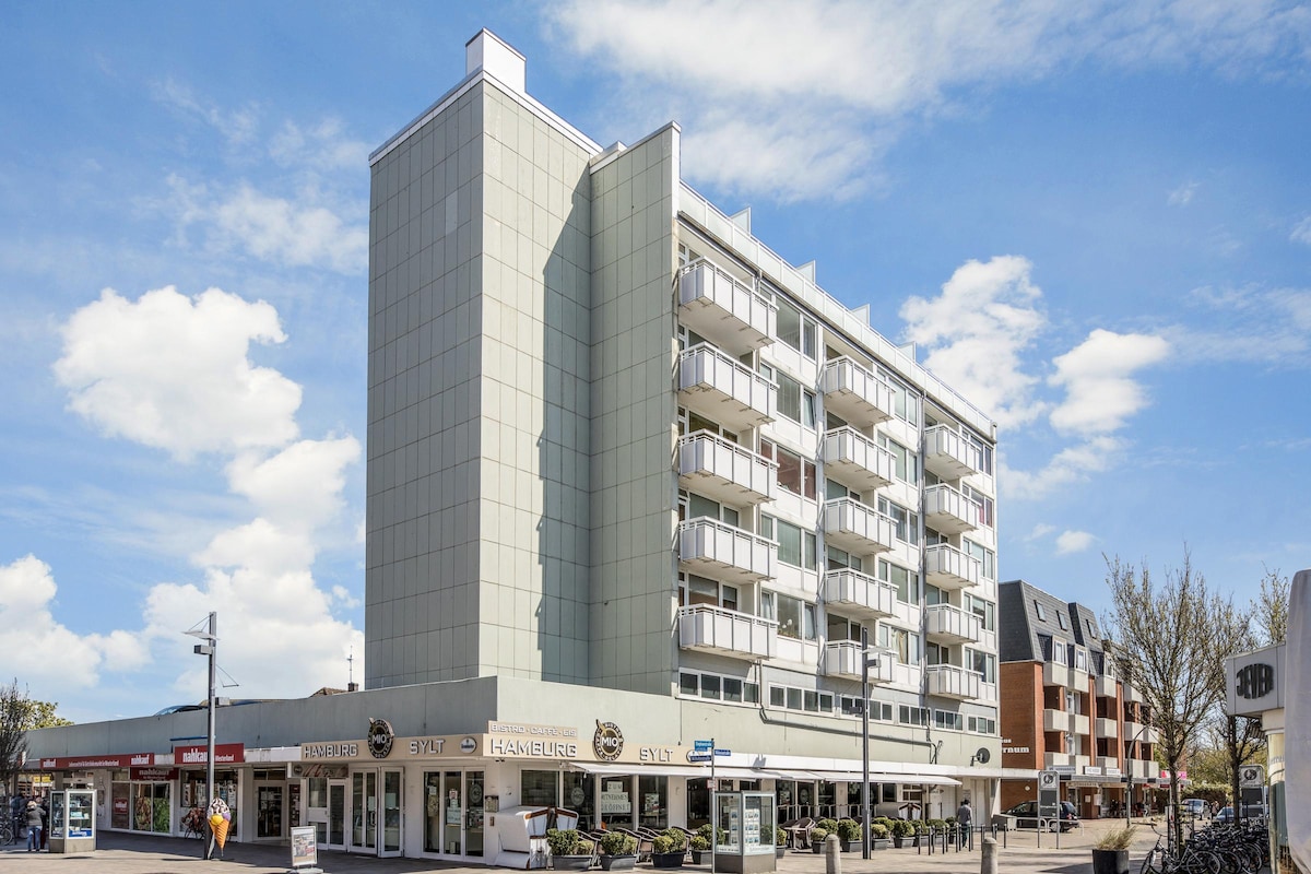 A modern five-story building is presented with a light-colored façade, featuring balconies on each floor. The entrance area includes café establishments, and the surrounding environment is lively with pedestrian pathways and landscaped greenery. The sky is clear with scattered clouds.