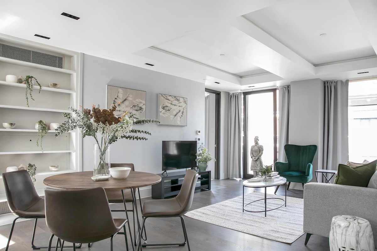 A modern living space features a round dining table surrounded by four chairs, offering a cozy setting. A plush green armchair and gray sofa invite relaxation, while light streams through large windows. Decorative shelves hold minimalist accents, adding to the contemporary aesthetic.