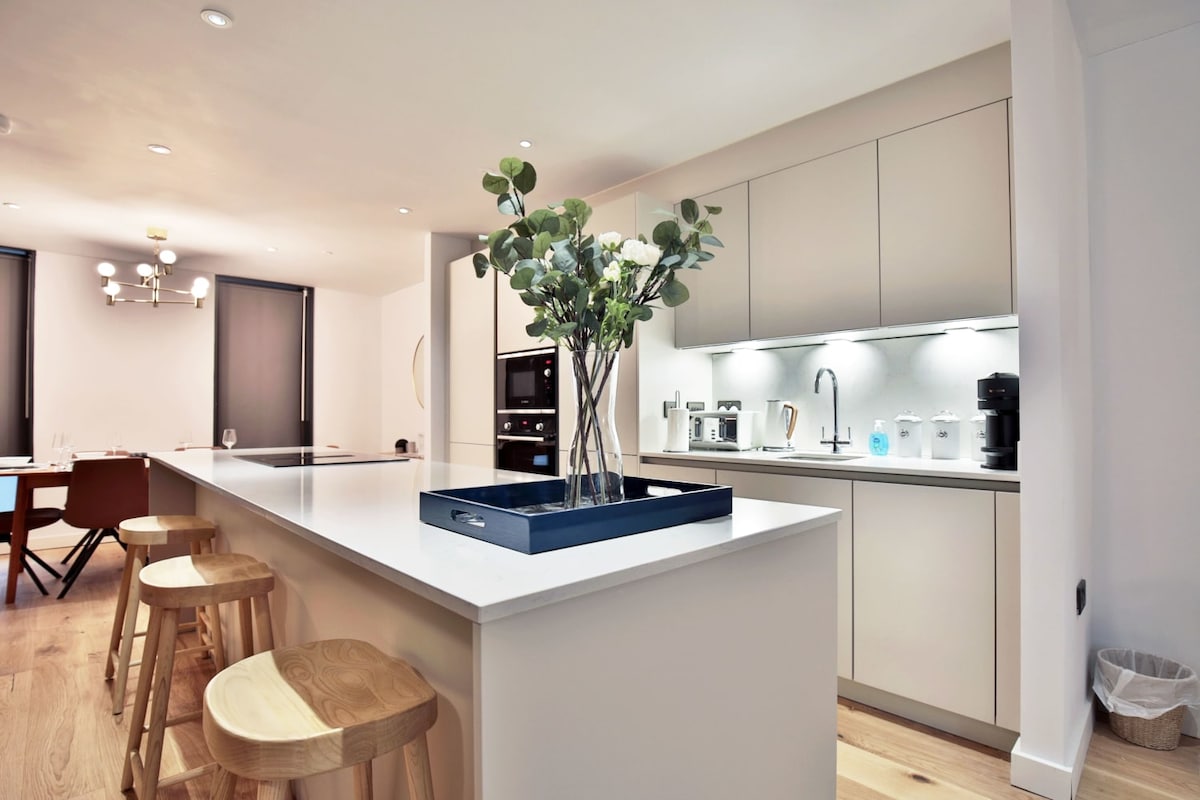 A modern kitchen is presented, featuring sleek cabinetry and an expansive island. The island is adorned with a decorative tray holding fresh greenery. Bar stools provide seating at one side, while under-cabinet lighting illuminates the workspace, highlighting the kitchen's contemporary design.