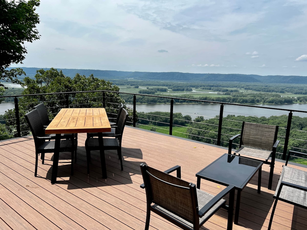 An outdoor deck features a wooden table surrounded by six black chairs, offering ample seating. Two lounge chairs and a glass-top table provide additional seating options. The deck overlooks expansive views of the Mississippi River and surrounding green hills under a partly cloudy sky.