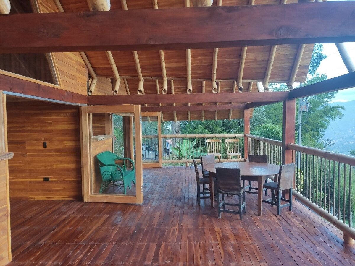 An outdoor deck features a large wooden table surrounded by multiple chairs, ideal for gatherings. A green armchair is seen in the corner. The roof showcases wooden beams, and expansive views of nature are visible in the background.
