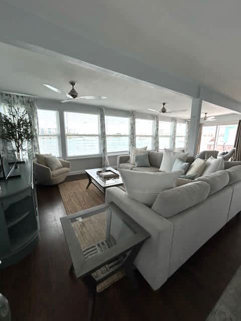 A bright living area features large windows that provide views of the water. Comfortable seating is arranged around a wooden coffee table. Light-colored furniture and subtle decorative elements create a relaxed, inviting space filled with natural light.