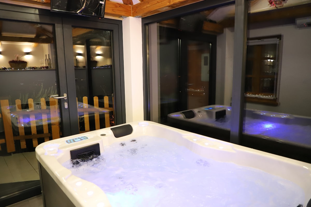 A relaxation area is highlighted by a hot tub with softly illuminated water, creating a calming ambiance. Large glass panels allow for views into the adjoining space, where subtle lighting reflects off the surfaces, enhancing the serene environment.