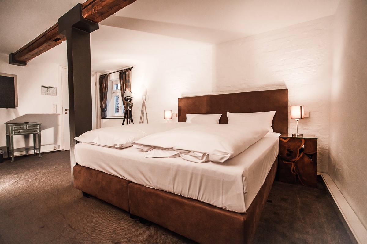 A spacious room features a large boxspring bed with crisp white linens. The wooden beam ceiling adds character, while soft lighting is provided by bedside lamps. A decorative tripod stands in the corner, contributing to the modern yet cozy atmosphere.