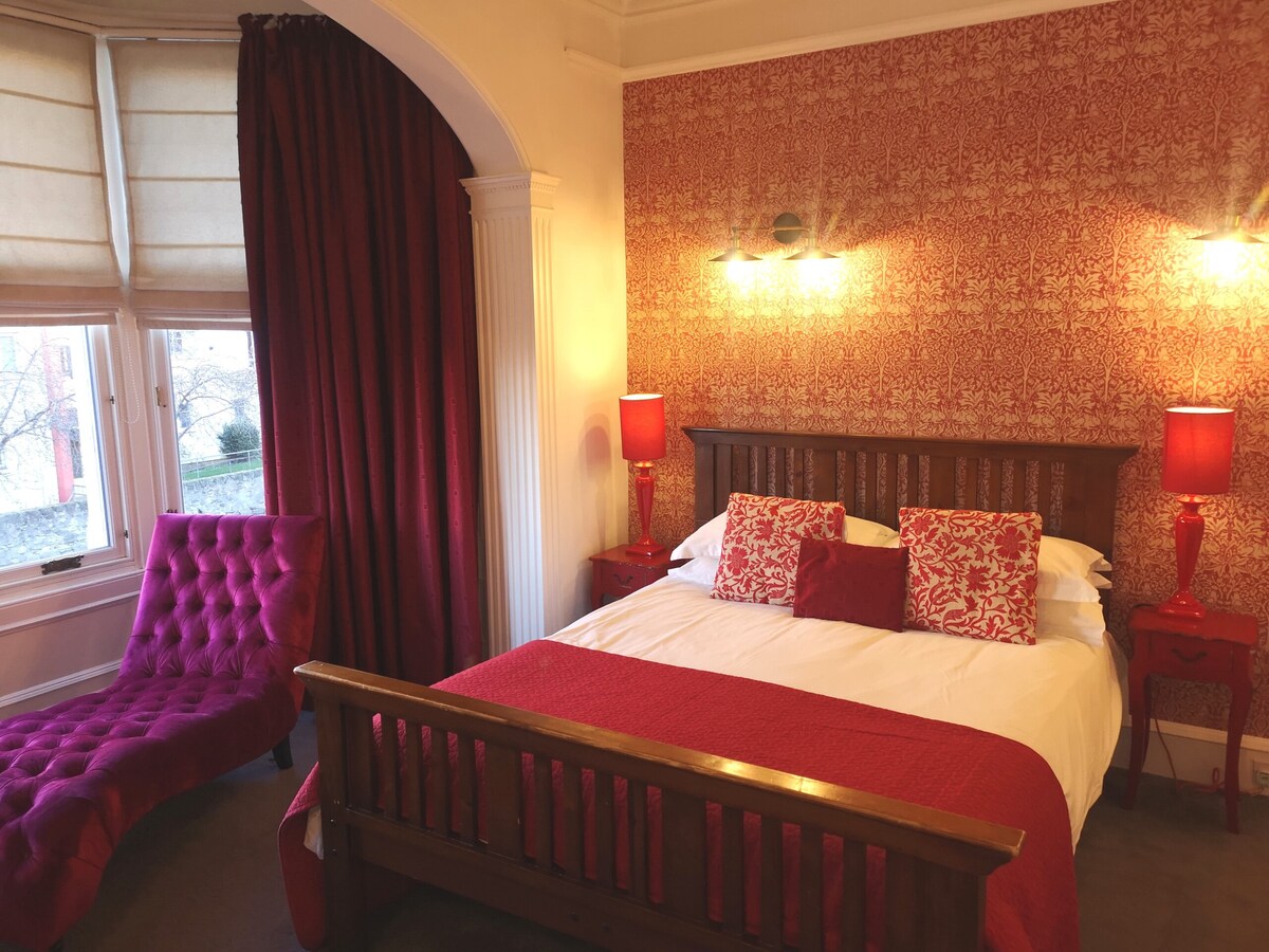 A cozy sleeping area features a wooden bed adorned with red and white bedding, complemented by decorative cushions. A plush chaise lounge in deep red sits beside the bed. Soft lighting is provided by bedside lamps against the patterned wallpaper and stylish curtain.