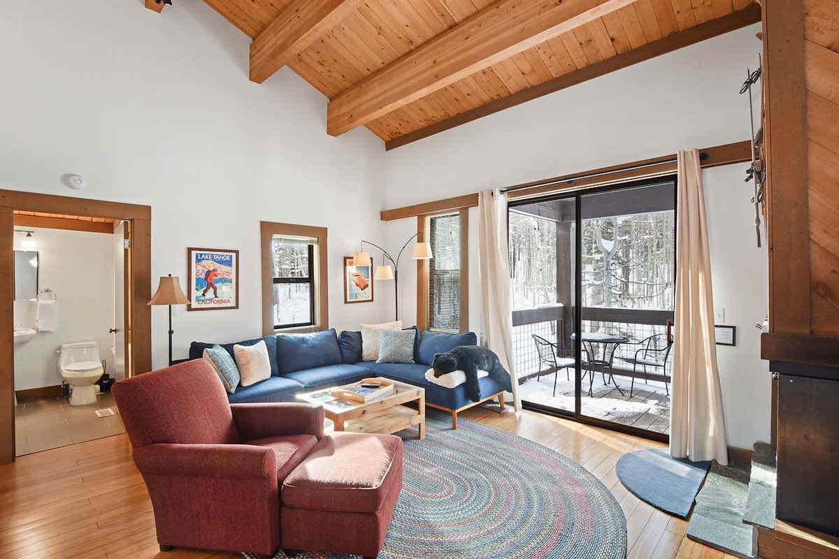 A cozy living area showcases a large sectional sofa with multiple decorative pillows, complemented by a coffee table and a soft area rug. A sliding glass door provides access to an outdoor space with a dining set, while warm wooden beams enhance the inviting atmosphere.