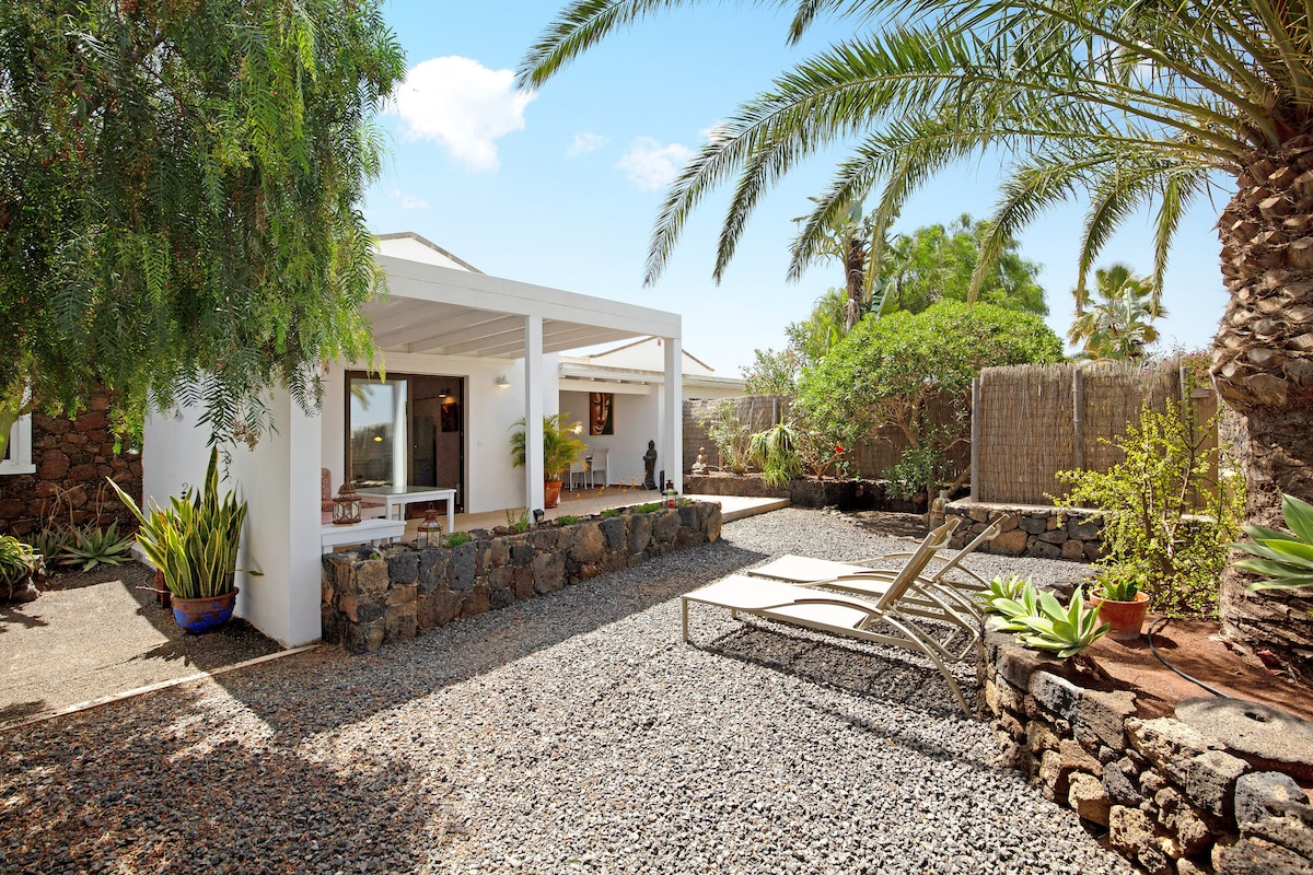 A private outdoor area is showcased, featuring a garden with tropical greenery and palm trees. A spacious covered terrace is visible, along with lounge chairs set on gravel. Natural stone walls delineate the area, providing a serene environment for relaxation.