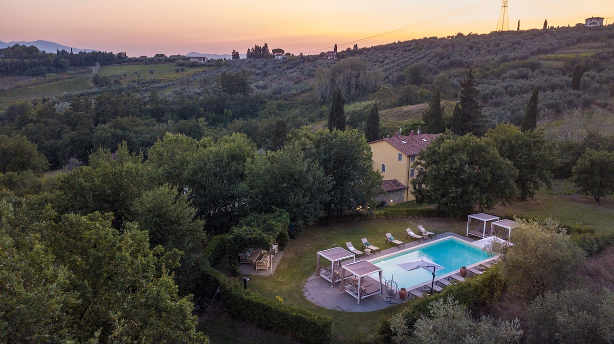 A tranquil scene captures a swimming pool surrounded by lush greenery and olive trees at sunset. Sun loungers are neatly arranged beside the pool, and shaded cabanas provide comfortable seating. The hillside in the background features a charming rural landscape, adding to the serene atmosphere.