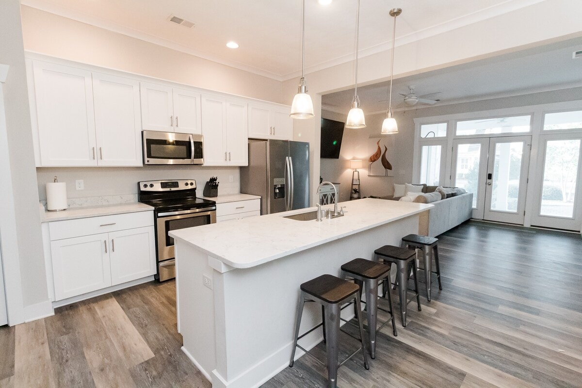 A modern kitchen is featured with white cabinetry, stainless steel appliances, and a spacious island with seating. Pendant lights hang above the island, illuminating the countertop. The open layout provides a view of the adjoining living area, with large windows inviting natural light.