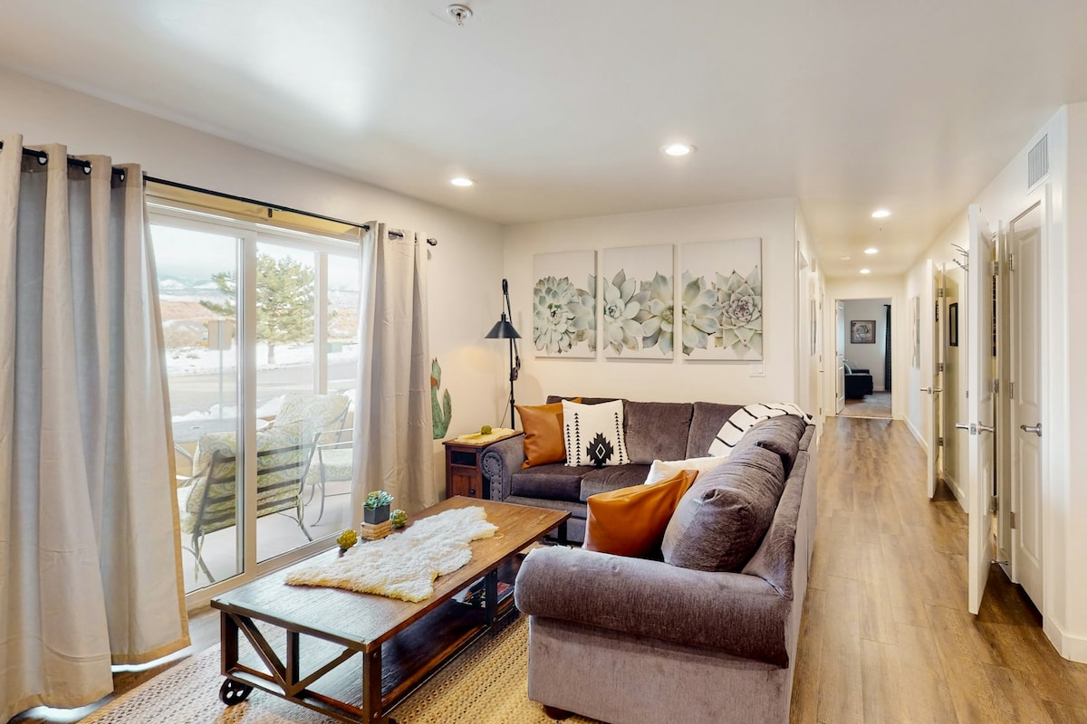 The living area features a comfortable gray sofa with decorative pillows. A wooden coffee table rests in front, complemented by a textured area rug. Large glass doors lead to an outdoor space, framed by light-colored curtains and showcasing wall artwork of succulent plants.