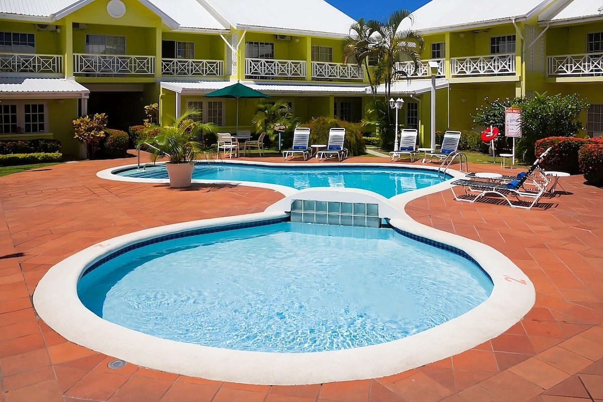 A curved swimming pool is surrounded by a sun deck with loungers and umbrellas. A smaller spa-like area is adjacent, with steps leading into the clear water. The vibrant green building in the background complements the tropical landscape.