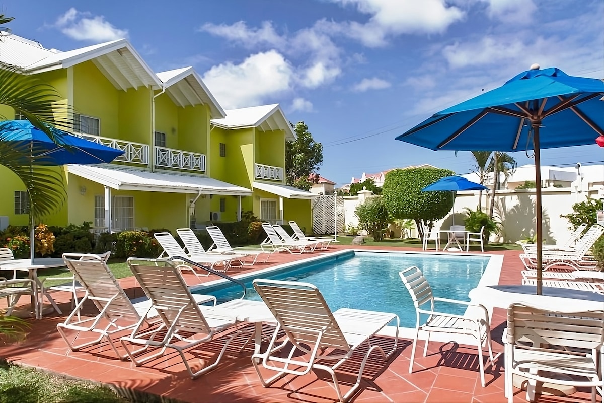 A welcoming outdoor pool area is surrounded by lounge chairs and colorful umbrellas. The vibrant yellow building serves as a backdrop, with lush greenery and flowering plants adding a touch of nature to the scene.