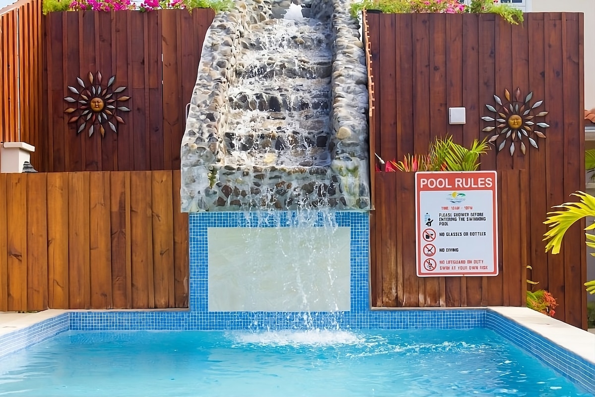 A refreshing swimming pool is highlighted, featuring a stone waterfall that cascades into the water. Surrounding the pool, wooden fencing adds warmth, complemented by vibrant floral accents. Clear signage detailing pool rules is visible nearby.