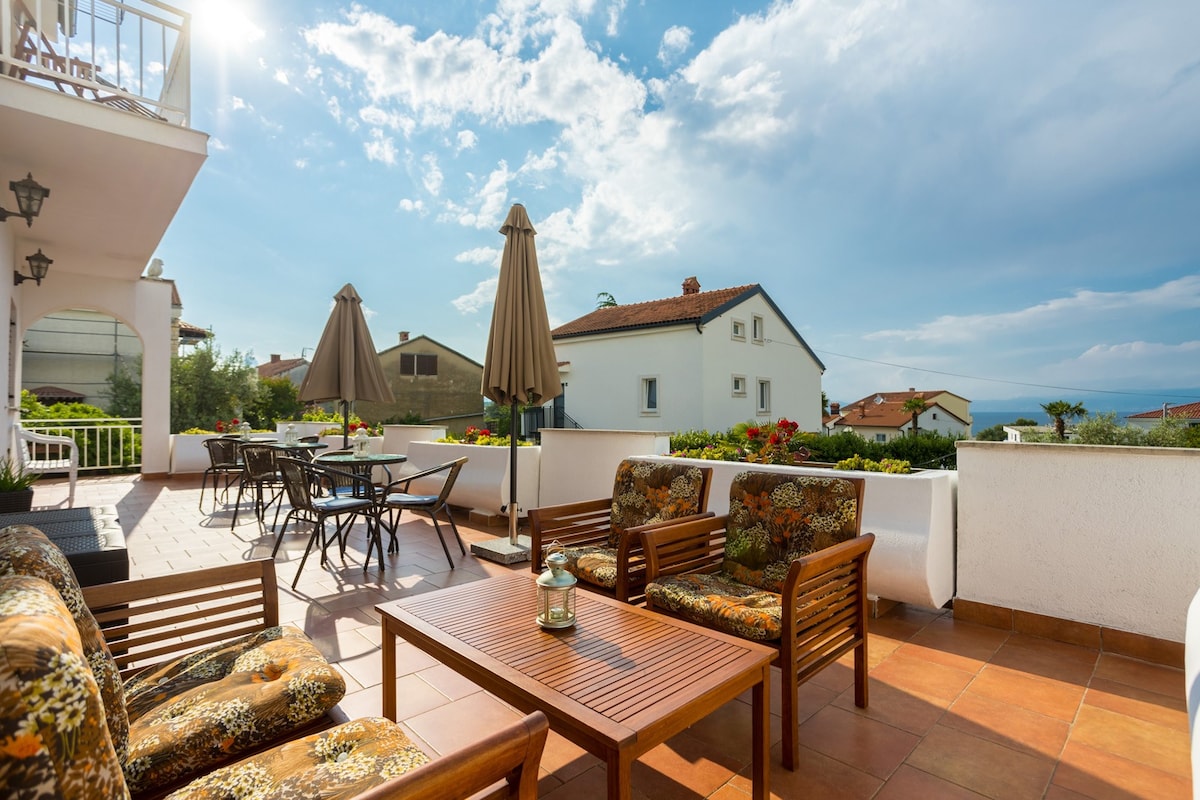 An inviting terrace is displayed, equipped with comfortable seating and a wooden table. Umbrellas provide shade over dining areas, surrounded by lush greenery. The backdrop features traditional houses and a clear blue sky, contributing to a welcoming outdoor environment.