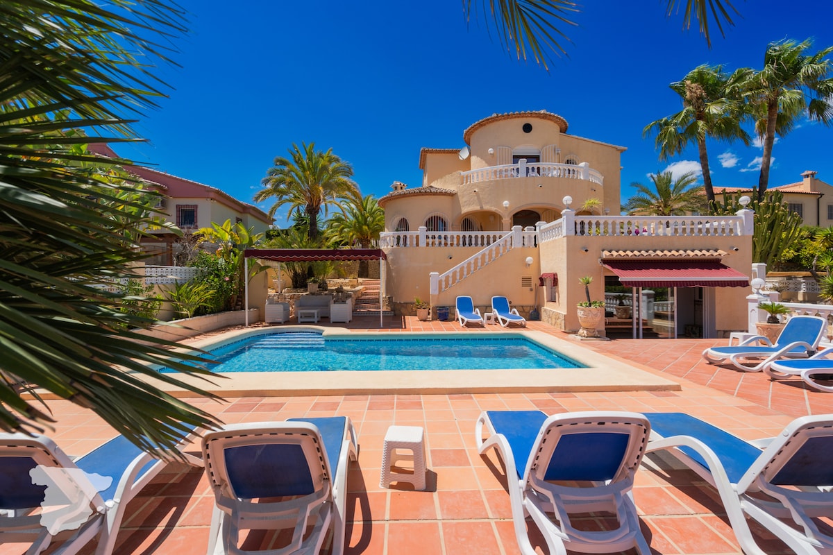 The exterior of the villa is highlighted, featuring a private swimming pool surrounded by sun loungers. Lush palm trees provide shade, and a terracotta tiled area offers space for relaxation. The villa’s multi-level structure showcases balconies and outdoor seating, conveying a welcoming atmosphere.
