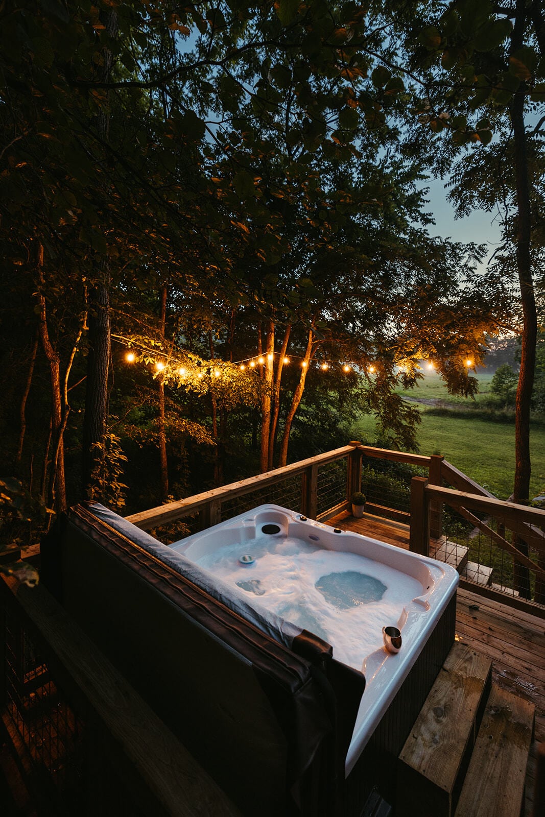 A hot tub is situated on a wooden deck, surrounded by a natural landscape. Soft lighting from string lights enhances the outdoor setting, while trees provide a sense of privacy. A gentle mist is seen in the distance, adding to the serene atmosphere.