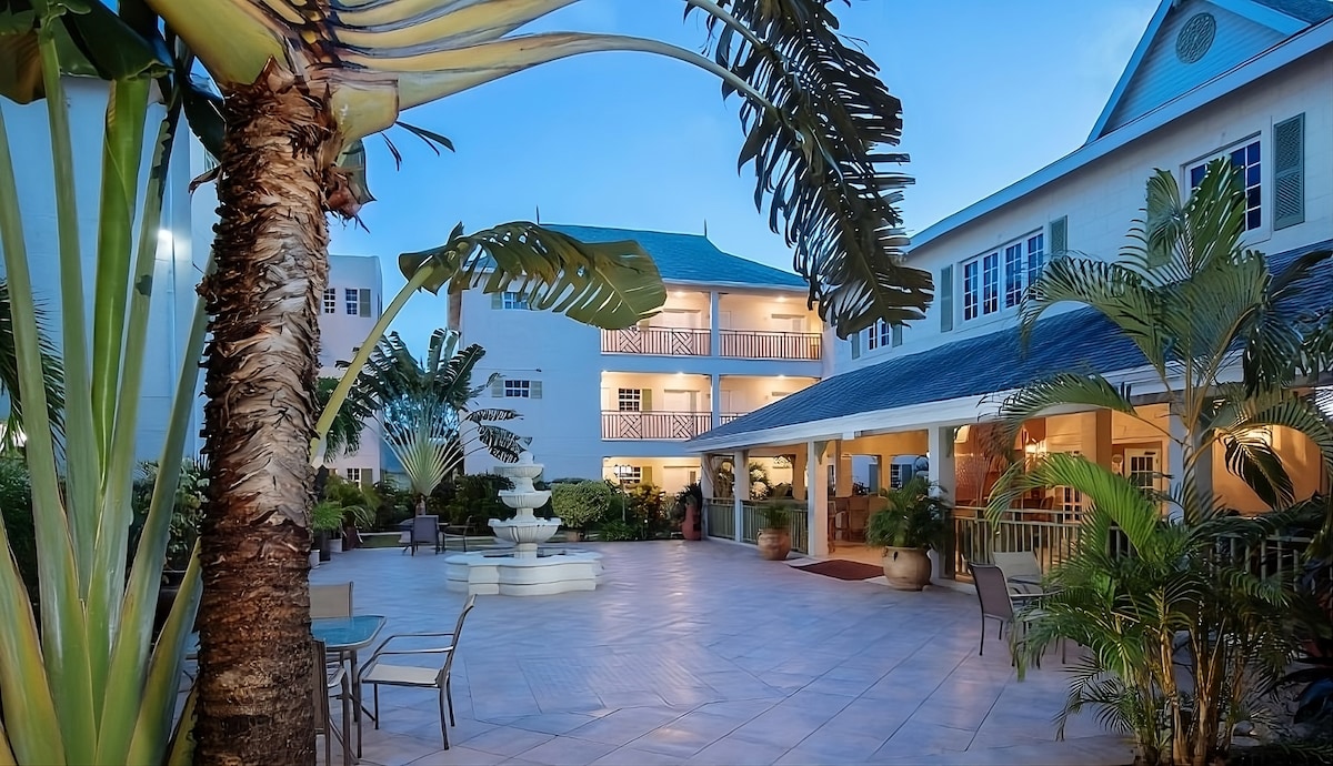 A serene courtyard is surrounded by lush tropical foliage and colorful architecture. A decorative fountain sits at the center, with seating areas and palm trees contributing to a relaxing atmosphere. The buildings feature multiple balconies overlooking the space, illuminated by soft evening light.
