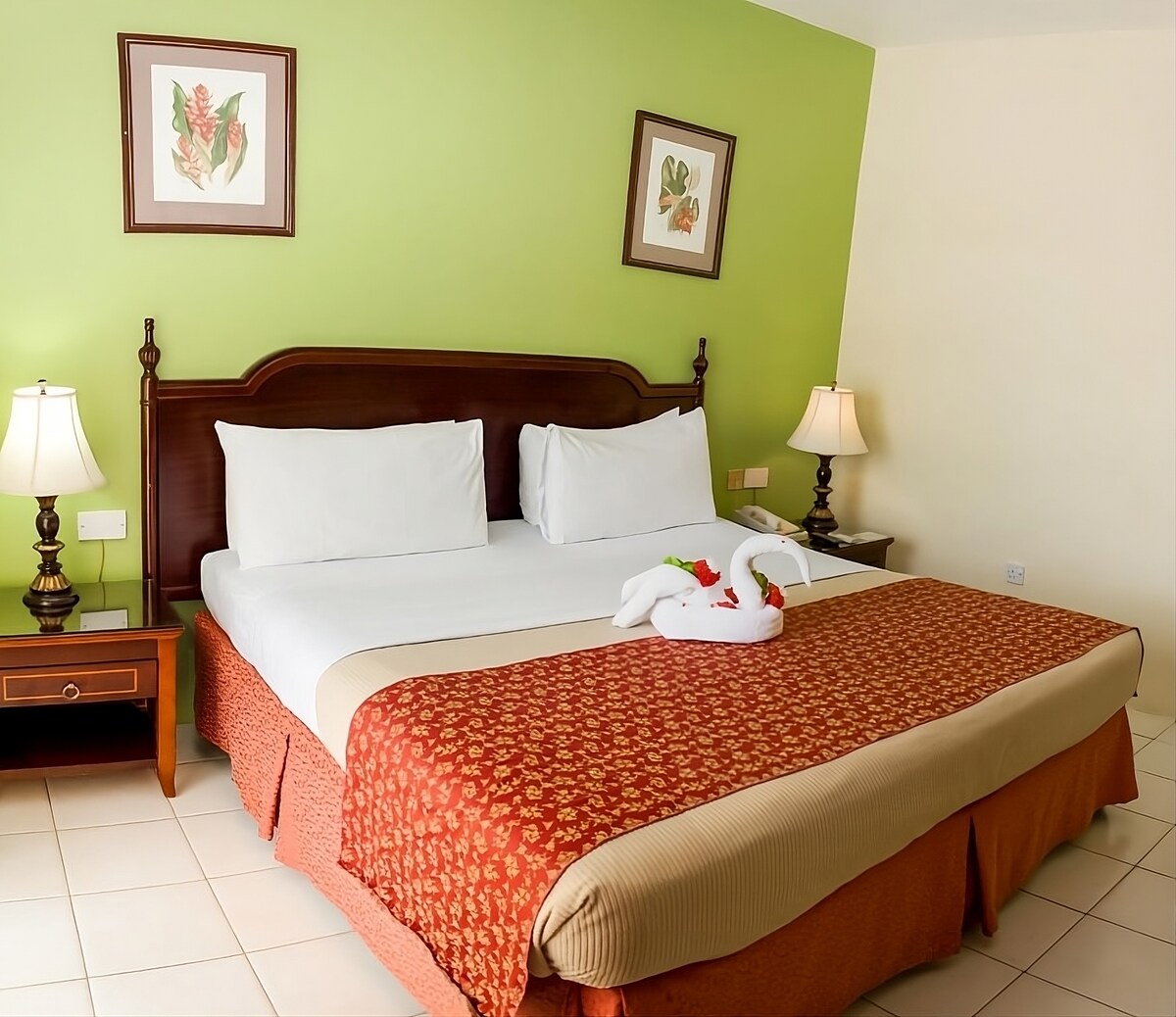 A spacious king room features a large bed with a decorative blanket, flanked by two bedside lamps. Green accent walls enhance the inviting atmosphere. Decorative artwork is displayed above the headboard, and soft, tiled flooring adds to the comfort of the space.