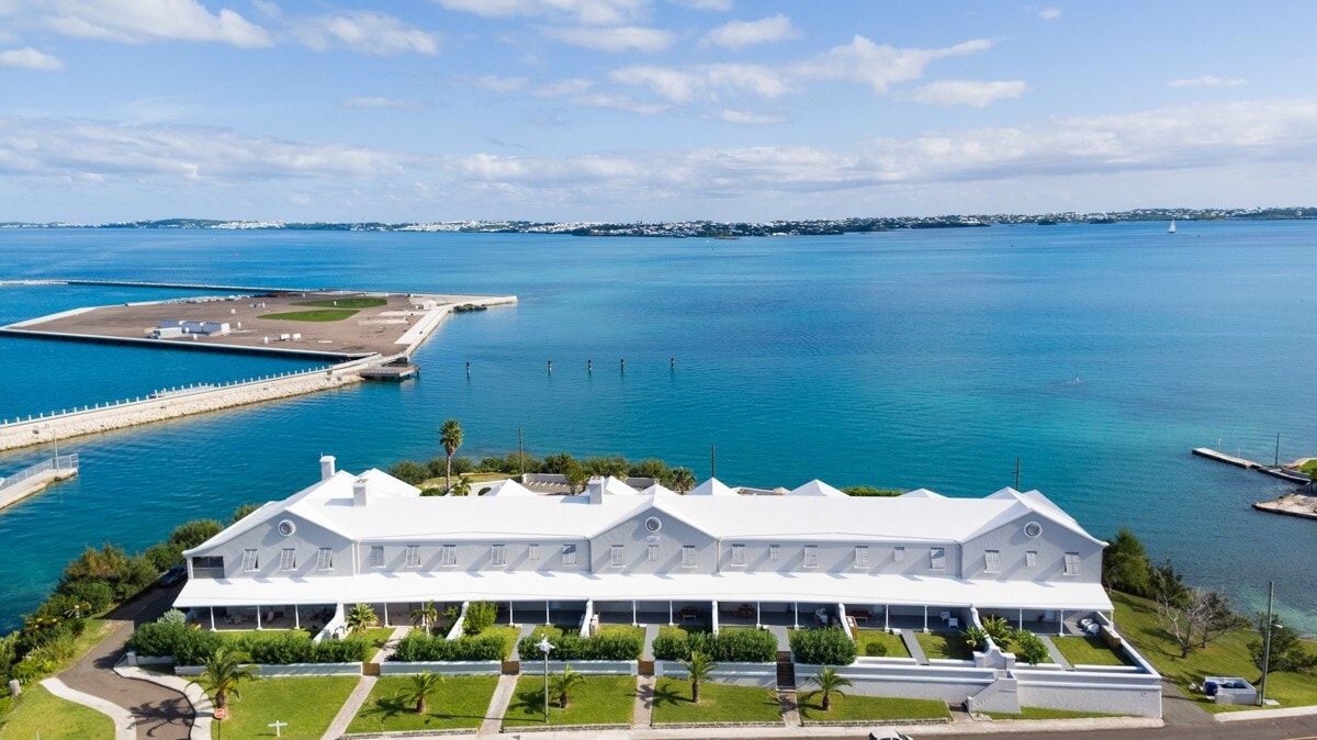 An aerial view of the upper apartment located on the water's edge is presented, showcasing a modern structure with a white exterior. The clear turquoise waters and the dock are visible in the background, enhancing the sense of tranquility and access to aquatic activities.