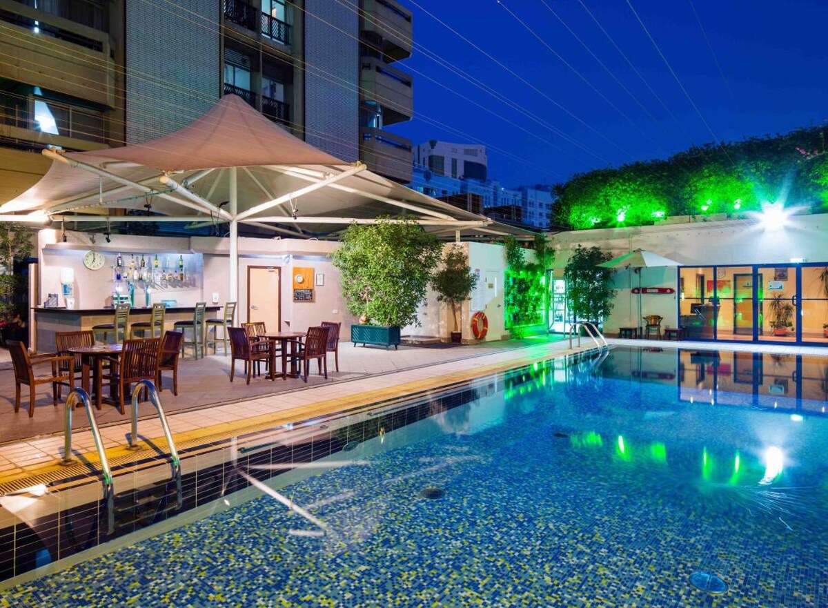 An outdoor pool area is surrounded by stylish seating and vibrant greenery, with a shaded bar featuring several tables. Ambient lighting reflects softly off the water's surface, creating a relaxing atmosphere during the evening.