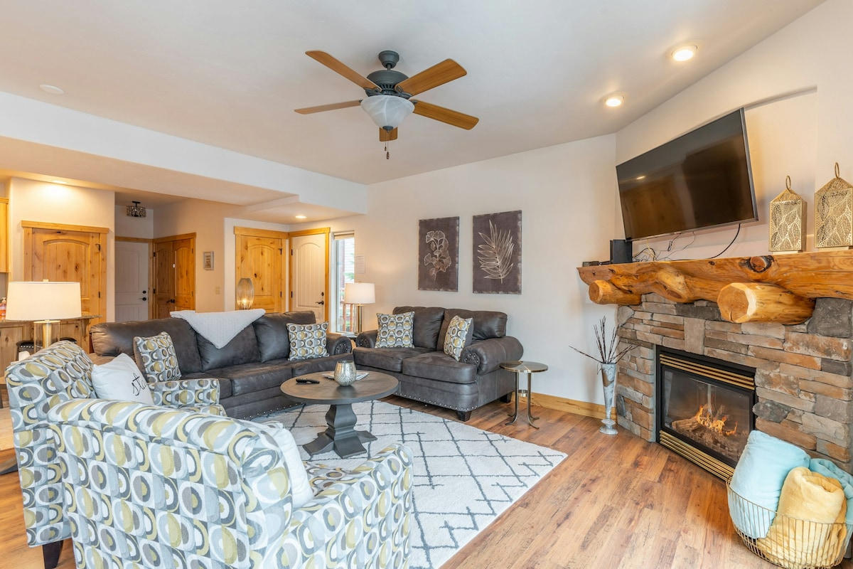 The spacious living area features a sectional sofa and coordinating accent chairs, arranged around a circular coffee table on a patterned area rug. A gas fireplace is framed by a stone mantle, and a wall-mounted TV is positioned above it, providing a cozy gathering space.