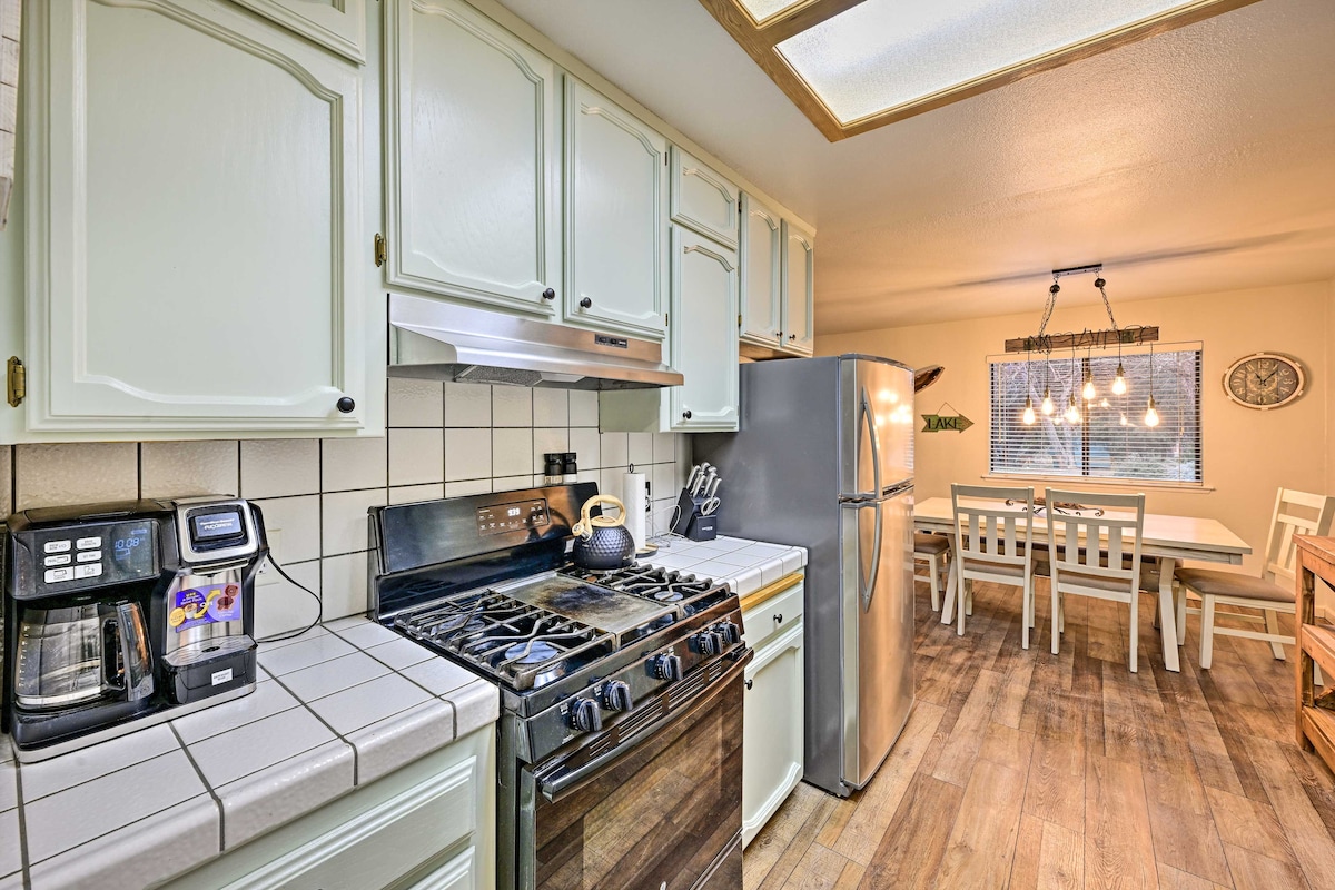 The kitchen features light-colored cabinetry and tiled countertops, with a gas stove and refrigerator visible. A coffee maker, toaster, and utensils are arranged on the counter. The dining area includes a light wood table and chairs, illuminated by pendant lights above.