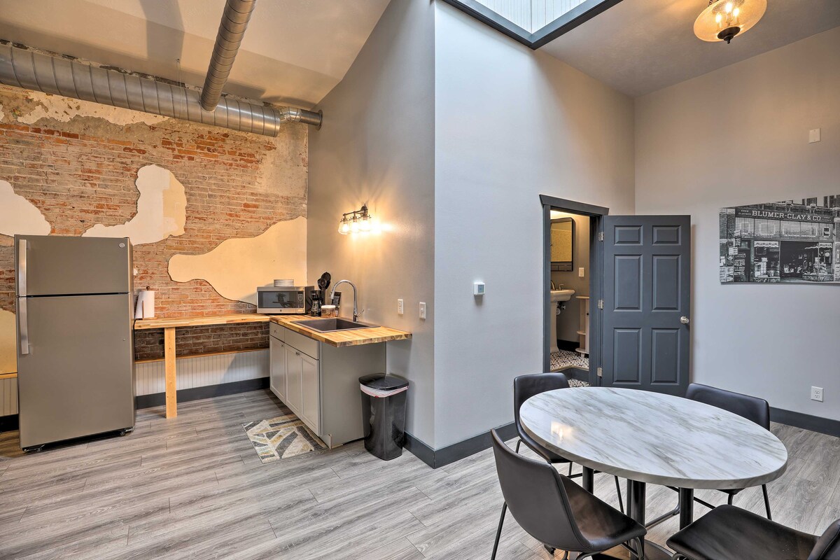 An open kitchen and dining area feature industrial-style design elements, with a brick accent wall, modern appliances, and a circular dining table. Large windows allow natural light to brighten the space, highlighting a combination of sleek countertops and wooden accents.
